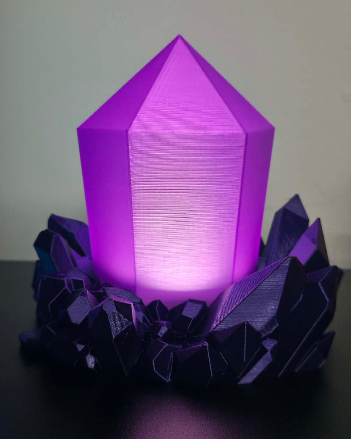 A purple 3D-printed crystal lamp glowing from within, resting on a dark, faceted base against a plain light gray background.